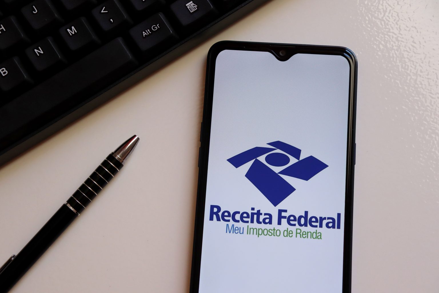 Imposto de Renda: tela de celular exibindo o aplicativo da Receita Federal, posicionada ao lado de um teclado e uma caneta.