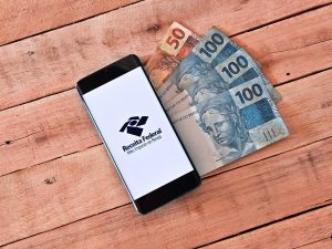 Aplicativo da Receita Federal aberto em um smartphone sobre mesa de madeira, ao lado de cédulas de 50 e 100 reais.