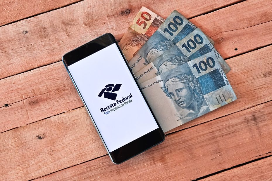 Aplicativo da Receita Federal aberto em um smartphone sobre mesa de madeira, ao lado de cédulas de 50 e 100 reais.