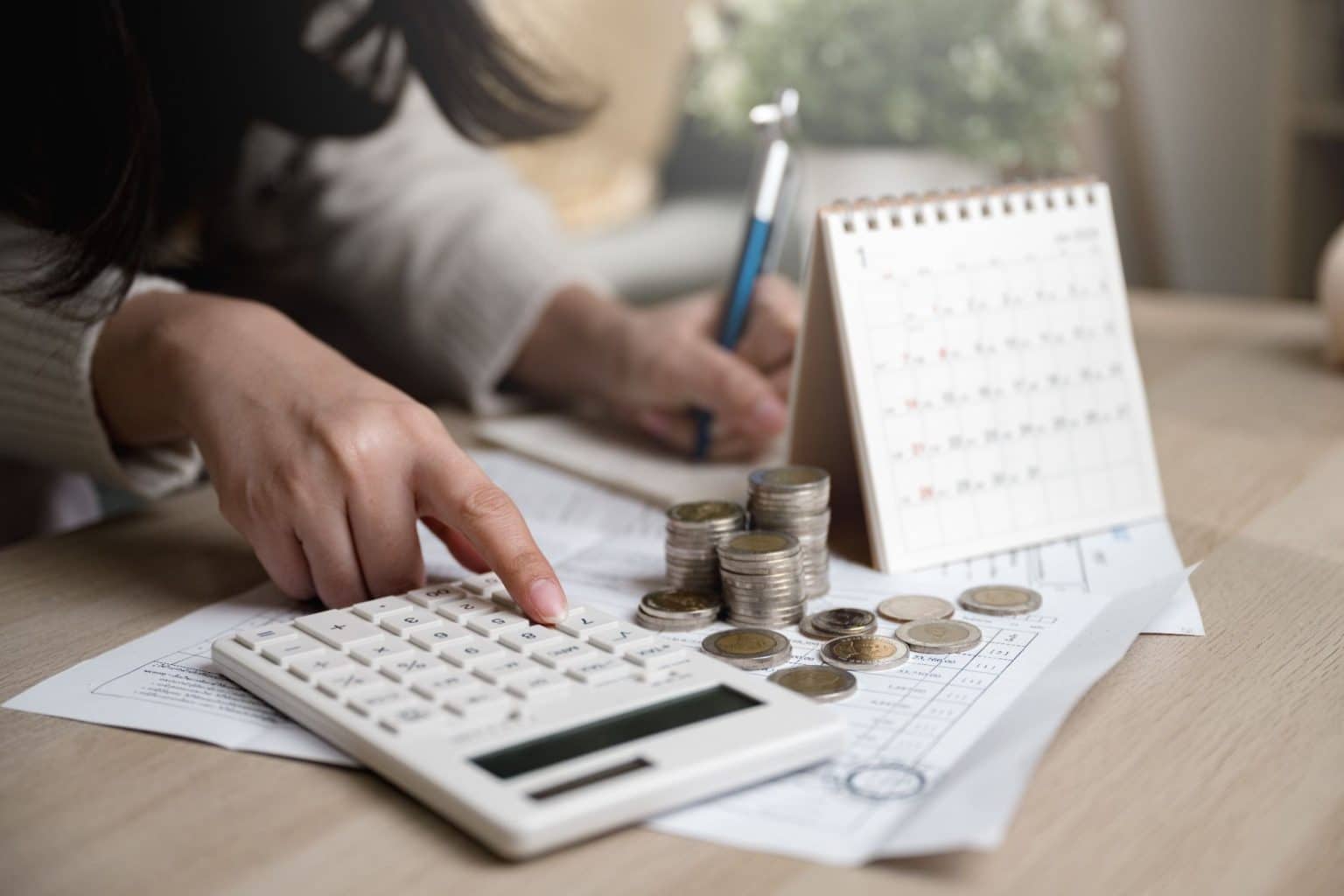 Pessoa calculando despesas com moedas e documentos sobre a mesa, usando calendário e calculadora para planejar como sair das dívidas rápido.