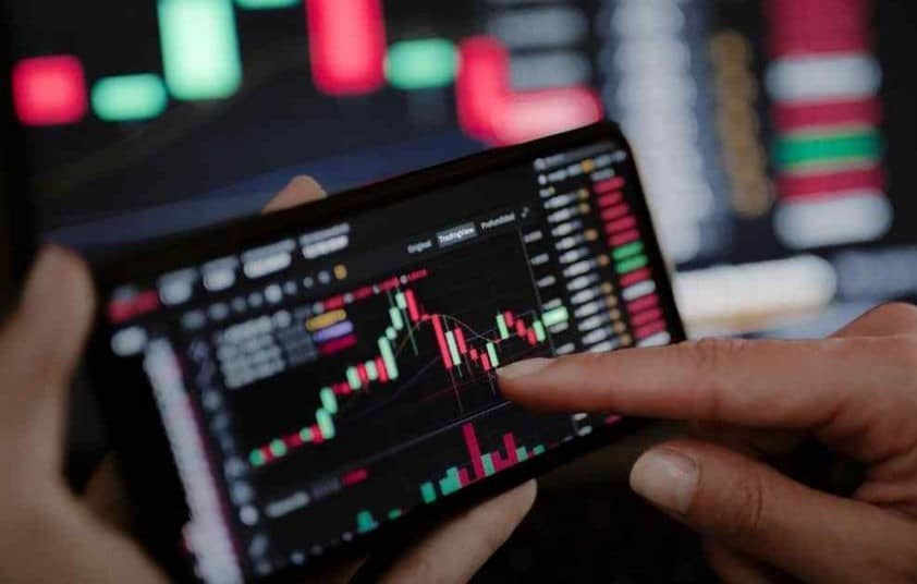 Pessoa analisa gráficos de ações em um celular, representando a volatilidade do mercado de renda variável e o risco de usar esse tipo de investimento para a reserva de emergência quanto guardar.