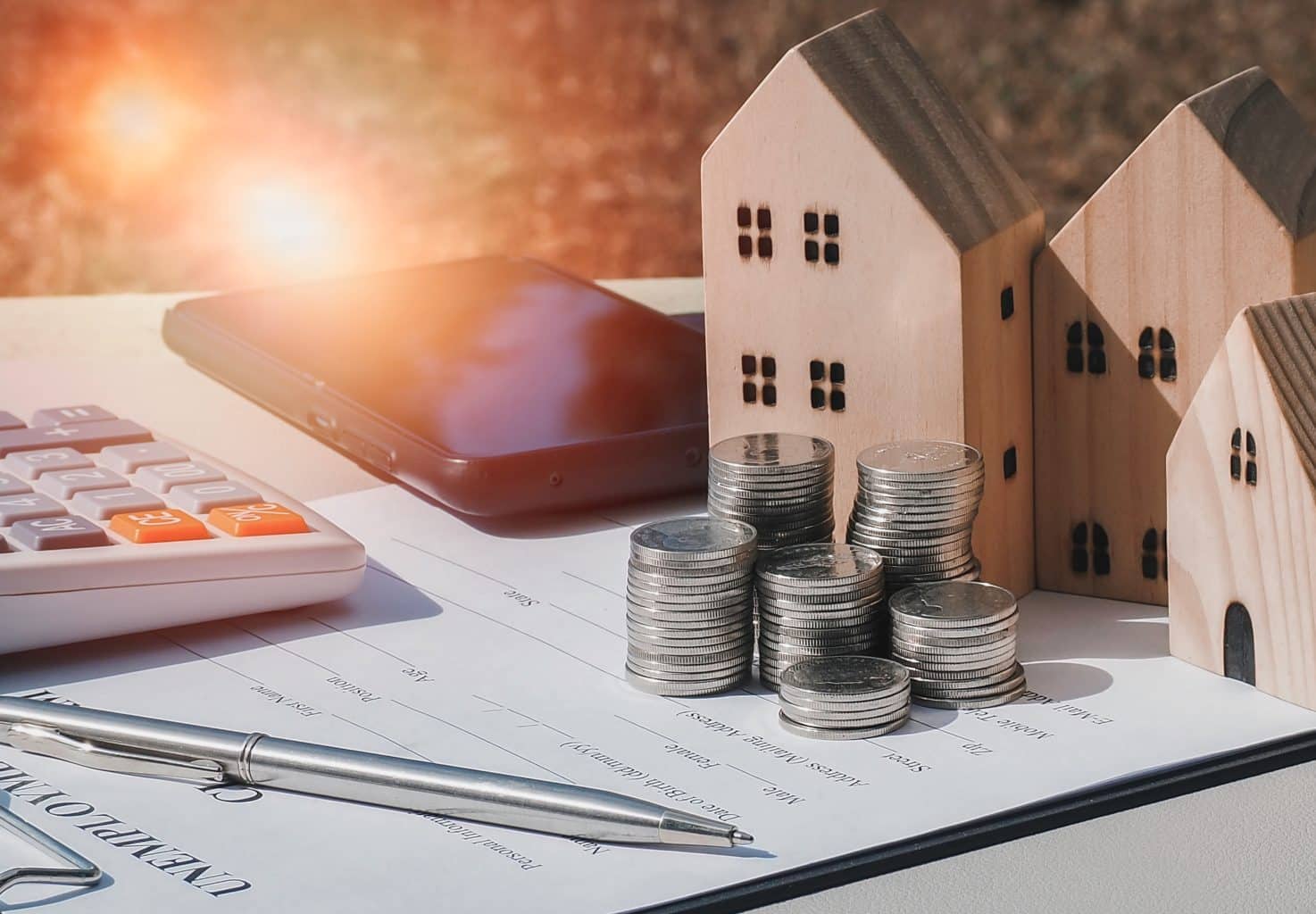 Pilhas de moedas ao lado de miniaturas de casas de madeira, calculadora e celular, representando planejamento financeiro e economia doméstica com dicas práticas.