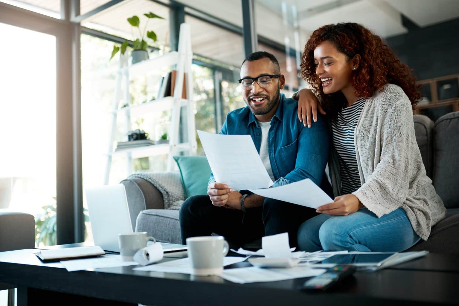 Casal sentado no sofá sorri enquanto analisa papéis e contas, representando um momento de controle financeiro familiar.