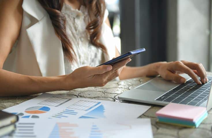 Mulher analisando gráficos financeiros no laptop enquanto usa o celular, simbolizando o uso de aplicativos para organizar finanças e manter o controle eficiente do orçamento pessoal.