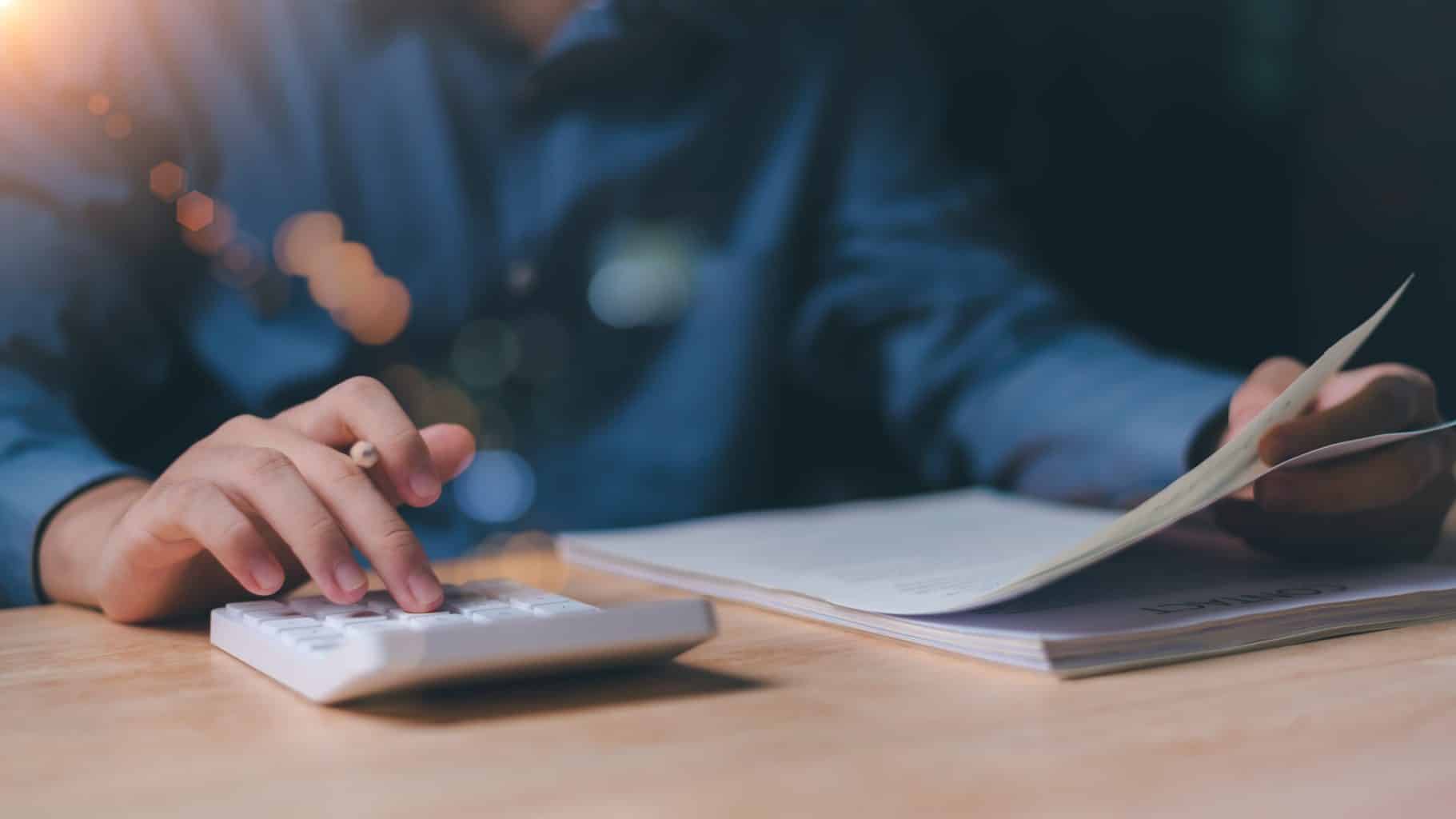 Pessoa analisando documentos e usando uma calculadora sobre uma mesa de madeira.