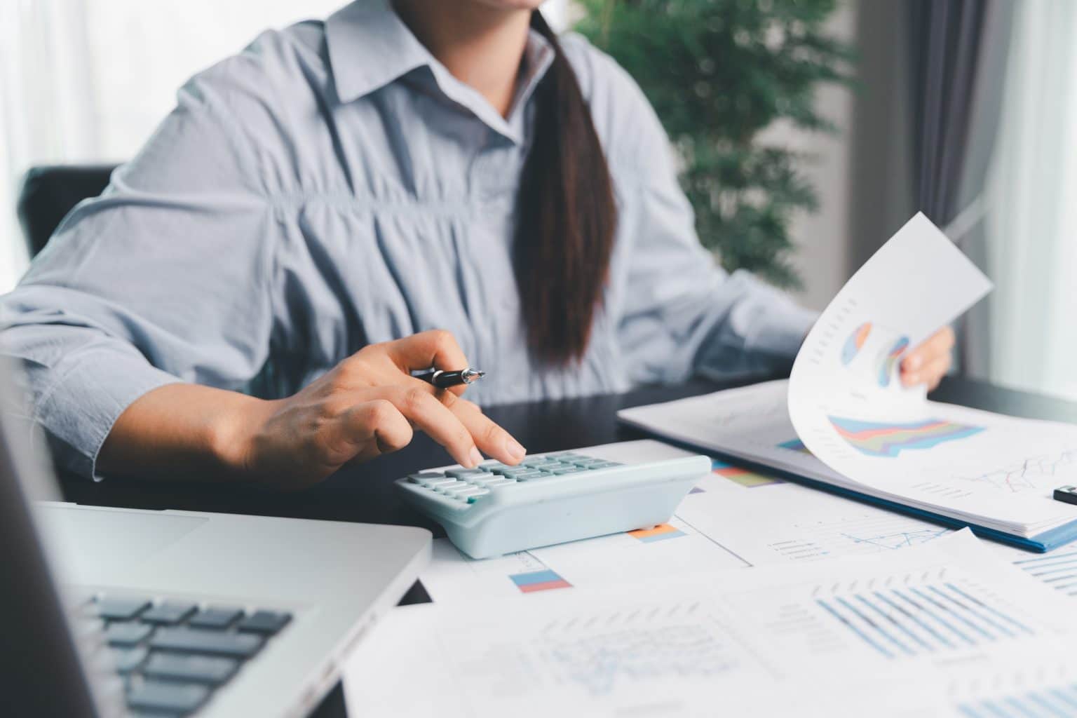 Mulher utilizando calculadora e analisando gráficos financeiros em ambiente de trabalho.