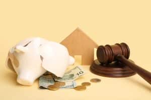 Cofrinho quebrado com dinheiro ao lado de martelo de juiz e miniatura de casa, simbolizando tarifa bancária e como evitar cobranças desnecessárias.