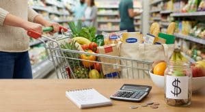 Carrinho de supermercado cheio de alimentos, com calculadora, lista de compras e moedas sobre a mesa, ilustrando como economizar nas compras de supermercado por meio do planejamento e controle de gastos.