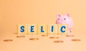 Imagem com a palavra “Selic” em blocos de madeira e um cofrinho ao fundo, representando a selic hoje e seu impacto nos investimentos e na economia.