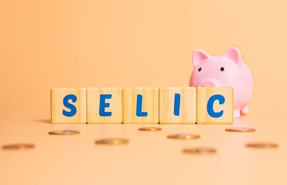 Imagem com a palavra “Selic” em blocos de madeira e um cofrinho ao fundo, representando a selic hoje e seu impacto nos investimentos e na economia.