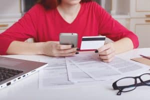 Pessoa sentada à mesa analisando contas e documentos enquanto segura um cartão de crédito e um celular, representando estratégias práticas de como reduzir a fatura do cartão de crédito e controlar gastos no dia a dia.